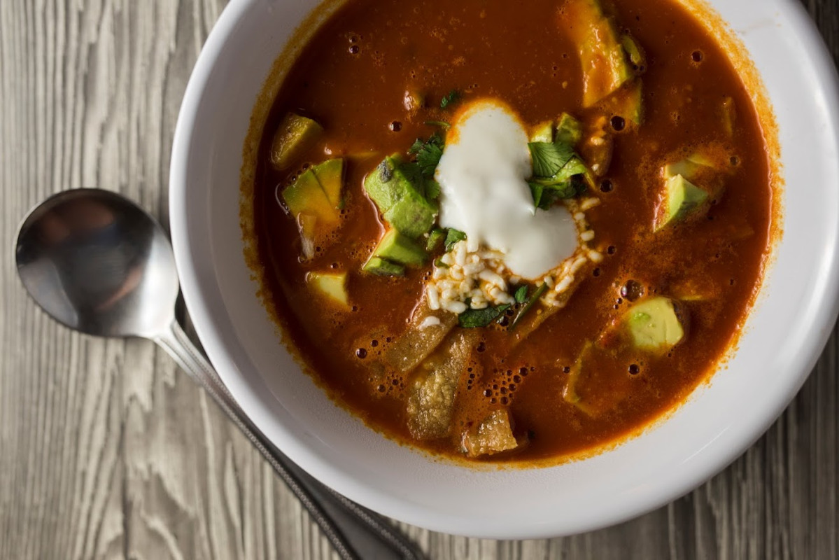 A warm tomato based soup served with slice of avocado, cheese, fried tortilla strips, and sour cream